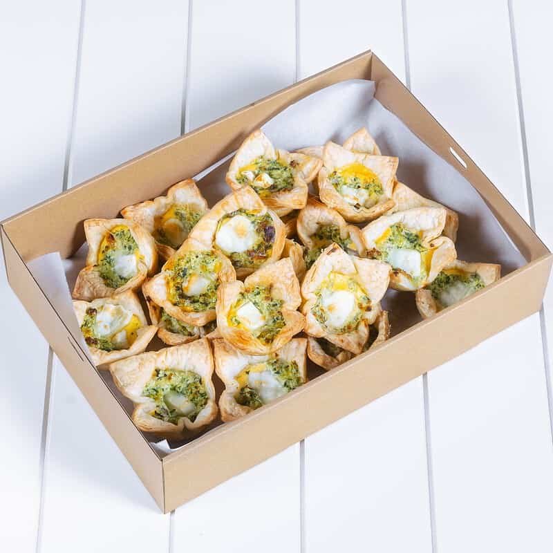 Mini pastry bites with green pesto and melted cheese in a cardboard box.