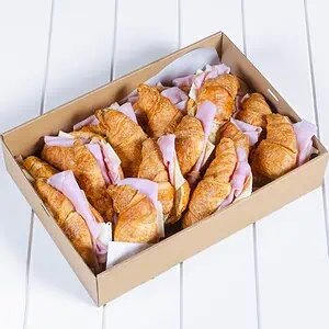 Box of croissant sandwiches with ham and cheese on white surface.