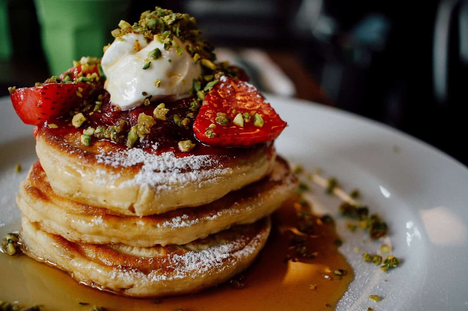 Stack of fluffy pancakes topped with strawberries, pistachios, and a dollop of cream.